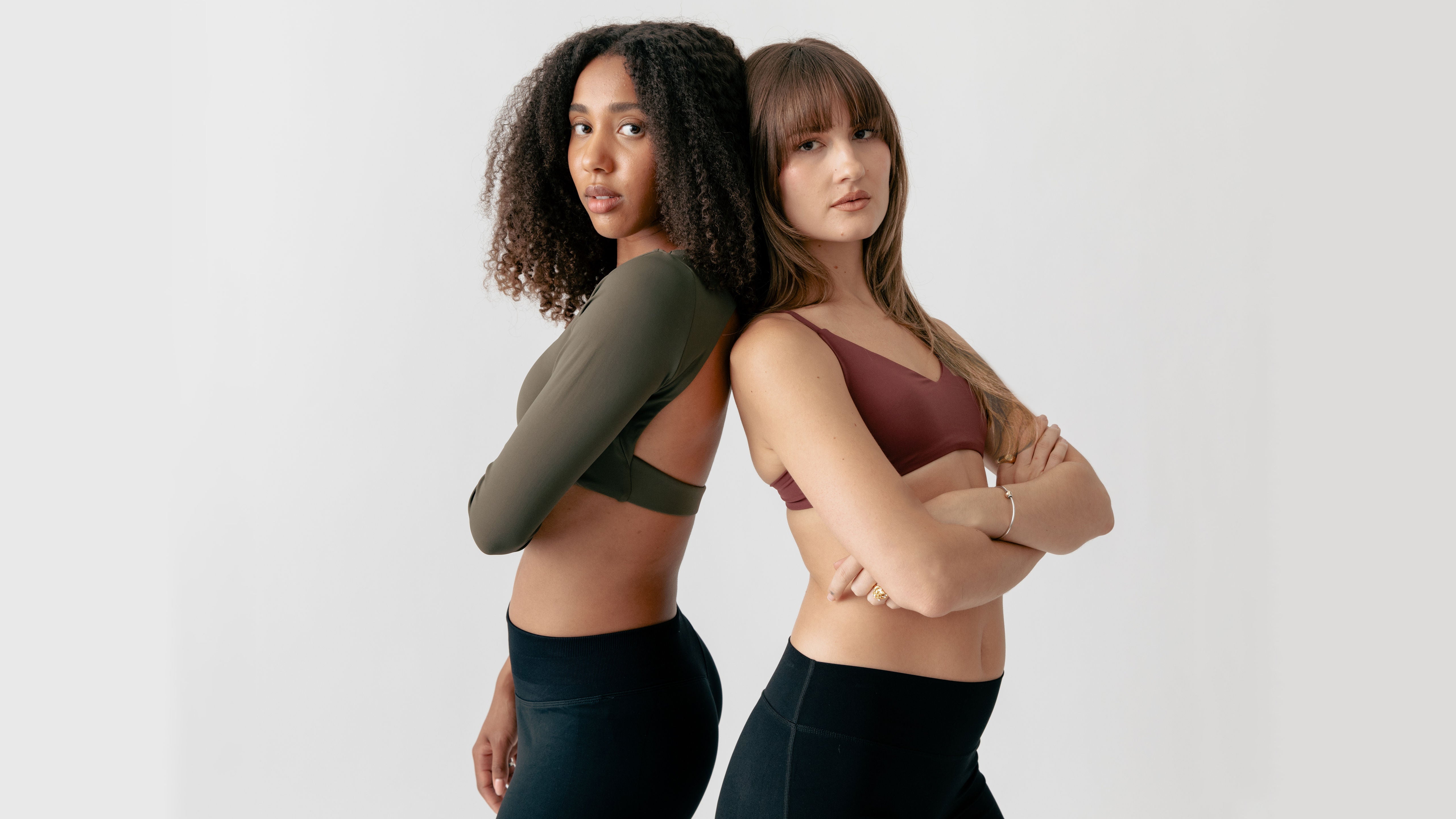 Two women wearing sports bras and leggings standing back-to-back against a white background