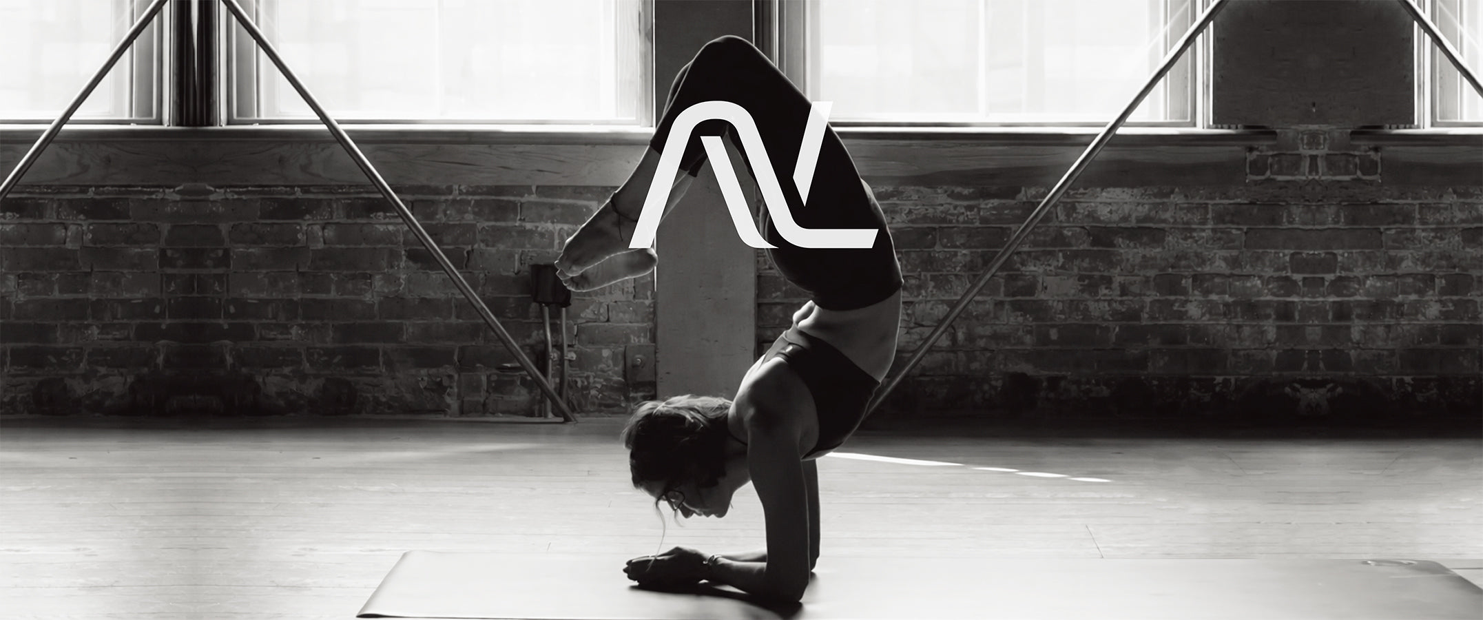 A black & white photograph of a woman performing a headstand in a studio with brick walls and windows with an NL logo on top.