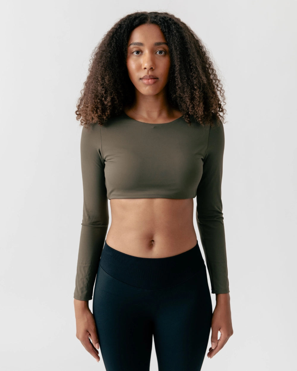 Woman wearing an olive green long-sleeve crop top and black leggings on a white background