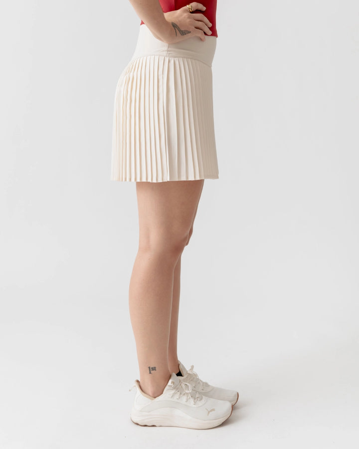 Person wearing a beige pleated skirt and white sneakers on a plain background