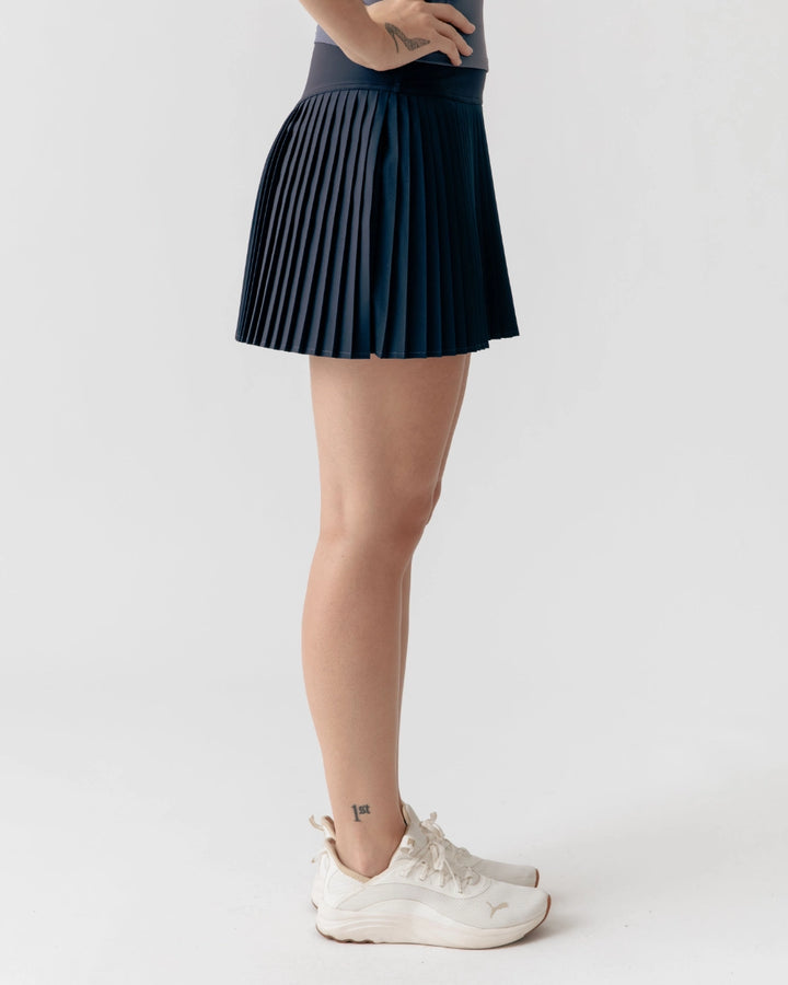 Person wearing a pleated black skirt and white sneakers on a plain background