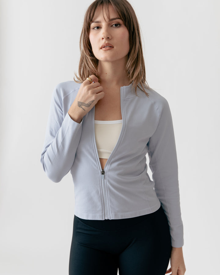 Woman wearing a light gray zip-up jacket over a white top and dark pants on a plain background