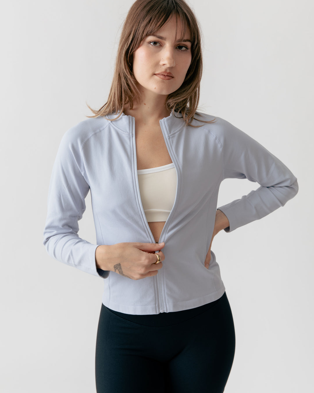 Woman wearing a light gray zip-up jacket over a white top and dark pants on a plain background