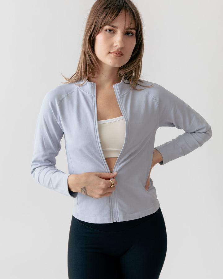 Woman wearing a light gray zip-up jacket over a white top and dark pants on a plain background