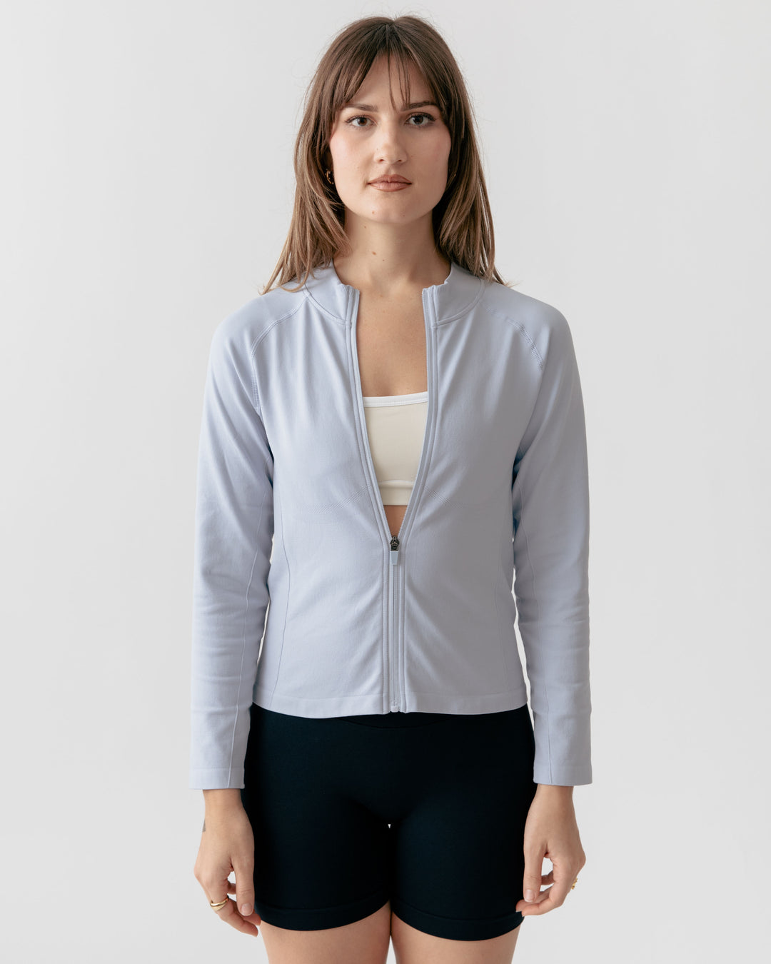 Woman wearing a light gray zip-up athletic jacket over a white sports bra and black shorts on a white background